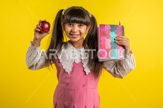بورتريه لطالبة عربية خليجية مبتمسة، ترتدي الزي المدرسي، تحمل الحقيبة المدرسية وبيدها الدفتر المدرسي وفاكهة التفاح الأحمر، فطور صباحي صحي، الإستعداد للعام الدراسي الجديد، التعليم الدراسي في السعودية، العودة إلى المدارس، خلفية صفراء