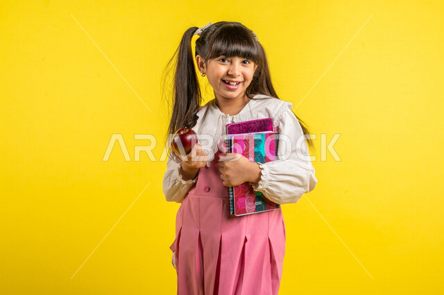 بورتريه لطالبة عربية خليجية مبتمسة، ترتدي الزي المدرسي، تحمل الحقيبة المدرسية وبيدها الدفتر المدرسي وفاكهة التفاح الأحمر، فطور صباحي صحي، الإستعداد للعام الدراسي الجديد، التعليم الدراسي في السعودية، العودة إلى المدارس، خلفية صفراء