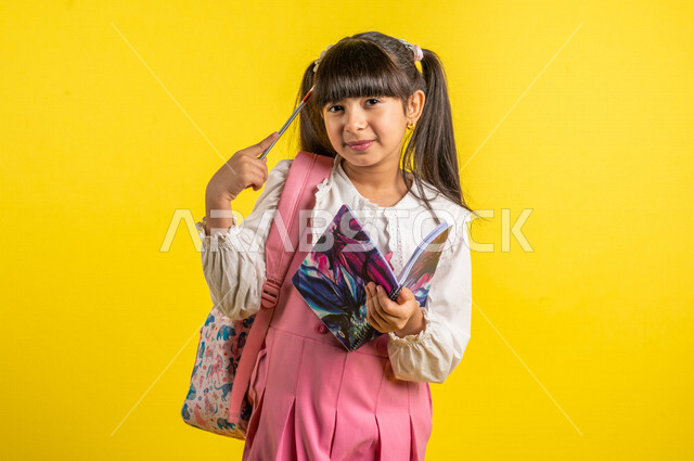 بورتريه لطالبة عربية خليجية مبتسمة، ترتدي الزي المدرسي، تحمل الحقيبة المدرسية وبيدها الدفتر المدرسي، تأدية الواجبات المدرسية، مراجعة الدروس والمناهج، الإستعداد للعام الدراسي الجديد، التعليم الدراسي في السعودية، العودة إلى المدارس، خلفية صفراء