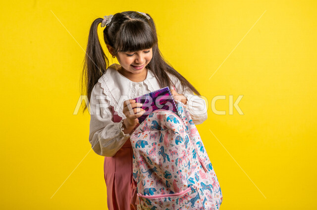 بورتريه لطالبة عربية خليجية مبتسمة، ترتدي الزي المدرسي، تحمل حقيبة الظهر المدرسية، إيماءات تدل على الحماس والسعادة لعودة المدارس، الإستعداد للعام الدراسي الجديد، التعليم الدراسي في المملكة العربية السعودية، خلفية صفراء، العودة إلى المدارس