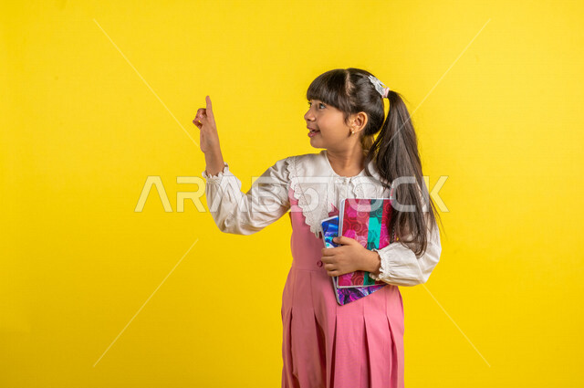 Portrait of a Saudi Arabian Gulf student, wearing school uniform, holding school notebooks, pointing with index finger at something, preparing for the new school year, school education in Saudi Arabia, yellow background, back to school