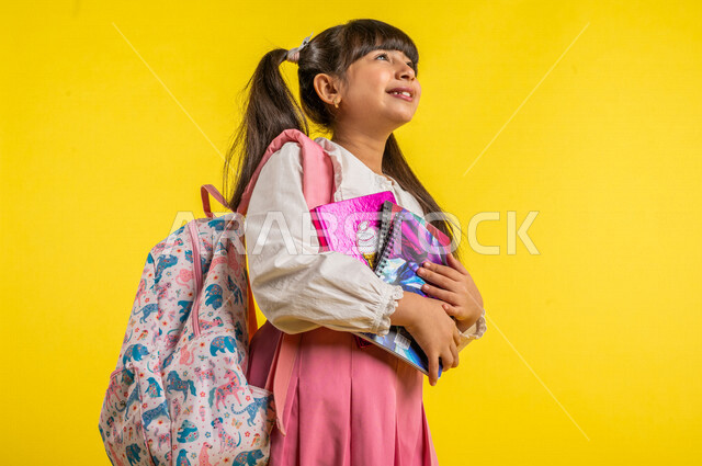 بورتريه لطالبة عربية خليجية مبتمسة، ترتدي الزي المدرسي، تحمل الحقيبة المدرسية وبيدها الدفاتر المدرسية، إيماءات تدل على السعادة لعودة المدارس، الإستعداد للعام الدراسي الجديد، التعليم الدراسي في السعودية، العودة إلى المدارس، خلفية صفراء