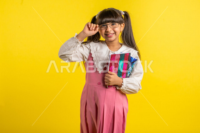 بورتريه لطالبة عربية خليجية مبتمسة، ترتدي الزي المدرسي، تحمل بيدها الدفاتر المدرسية، إيماءات تدل على السعادة لعودة المدارس، الإستعداد للعام الدراسي الجديد، التعليم الدراسي في السعودية، العودة إلى المدارس، خلفية صفراء