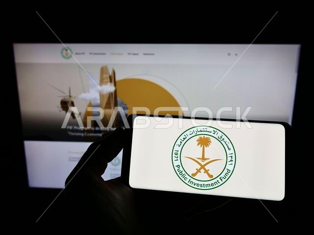 شخص يحمل هاتفًا ذكيًا عليه شعار صندوق الاستثمارات العامة السعودي (PIF) على الشاشة أمام موقع إلكتروني. التركيز على شاشة الهاتف.
