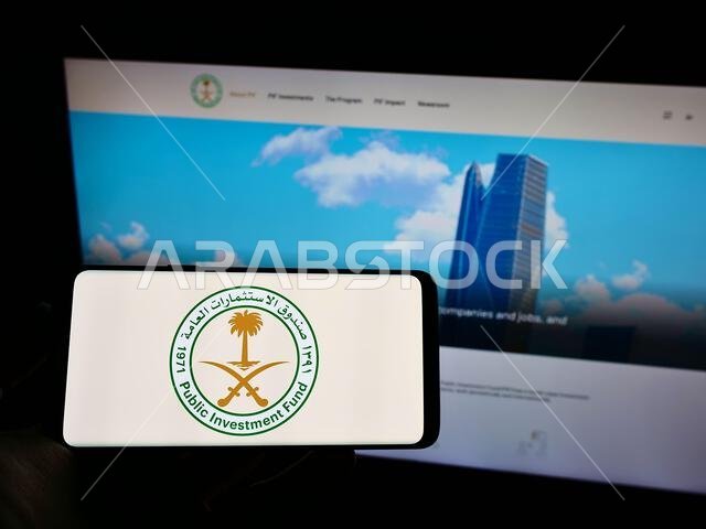 Person holding mobile phone with logo of Saudi Arabian Public Investment Fund (PIF) on screen in front of web page. Focus on phone display.