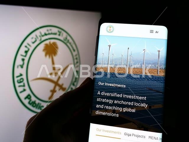شخص يحمل هاتفًا ذكيًا يعرض صفحة ويب لصندوق الاستثمارات العامة السعودي (PIF) أمام الشعار. التركيز على منتصف شاشة الهاتف. شخص يحمل هاتفًا ذكيًا يعرض صفحة ويب لصندوق الاستثمارات العامة السعودي (PIF) أمام الشعار. التركيز على منتصف شاشة الهاتف.