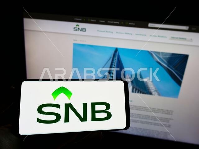 Person holding cellphone with logo of financial company Saudi National Bank (SNB) on screen in front of business webpage. Focus on phone display.