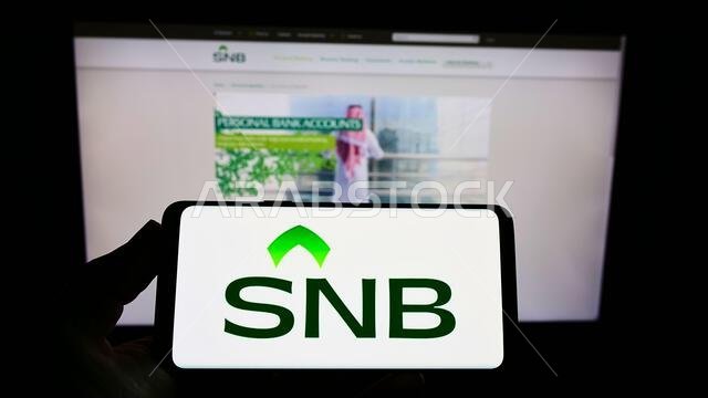Person holding smartphone with logo of financial company Saudi National Bank (SNB) on screen in front of website. Focus on phone display.