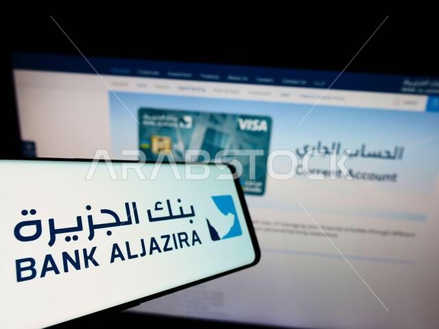 هاتف ذكي يحمل شعار بنك الجزيرة السعودي (BAJ) على شاشته أمام موقع إلكتروني خاص بالبنك. التركيز على منتصف يسار شاشة الهاتف.
