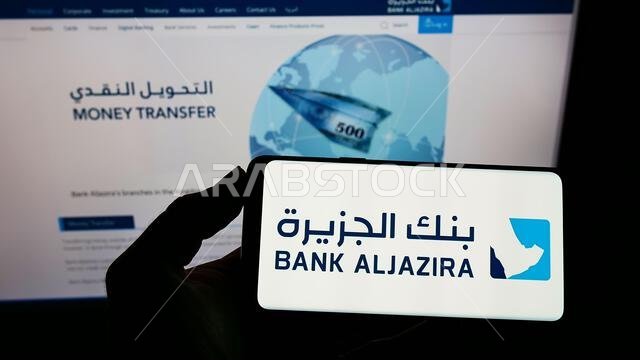 شخص يحمل هاتفًا محمولًا عليه شعار شركة بنك الجزيرة (BAJ) السعودية على الشاشة أمام صفحة الويب الخاصة بالشركة. التركيز على شاشة الهاتف.