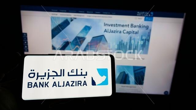 شخص يحمل هاتفًا ذكيًا عليه شعار شركة بنك الجزيرة (BAJ) السعودية على الشاشة أمام موقع إلكتروني. التركيز على شاشة الهاتف.