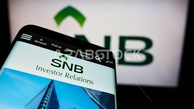 Mobile phone with logo of financial company Saudi National Bank (SNB) on screen in front of business webpage. Focus on phone display.