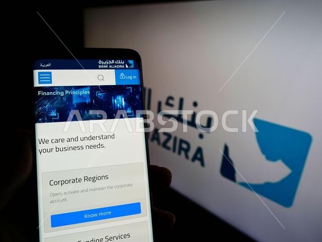 شخص يحمل هاتفًا محمولًا يعرض موقع بنك الجزيرة (BAJ) السعودي على الشاشة أمام الشعار. التركيز على منتصف شاشة الهاتف.