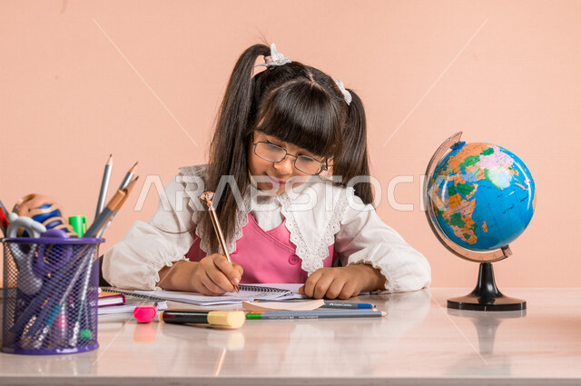 Portrait of a Saudi Arabian Gulf student, in school uniform, sitting at a table, next to a group of pens, textbooks and a globe, doing homework, preparing for the new school year, school education in Saudi Arabia, back to school, colorful background