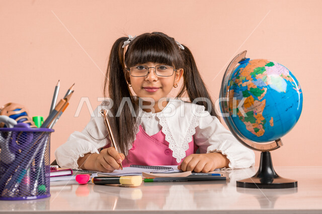 Portrait of a Saudi Arabian Gulf student, in school uniform, sitting at a table, next to a group of pens, textbooks and a globe, doing homework, preparing for the new school year, school education in Saudi Arabia, back to school, colorful background