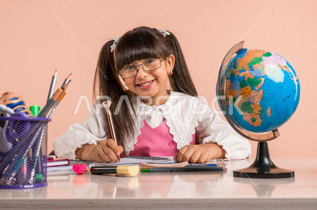 Portrait of a Saudi Arabian Gulf student, in school uniform, sitting at a table, next to a group of pens, textbooks and a globe, doing homework, preparing for the new school year, school education in Saudi Arabia, back to school, colorful background