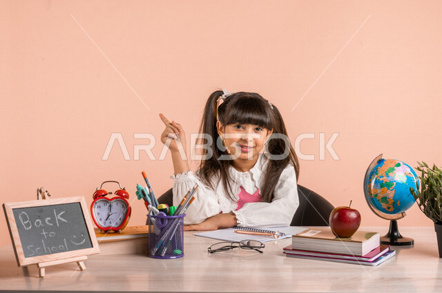 Portrait of a Saudi Arabian Gulf student, in school uniform, sitting at the table, next to a group of pens, textbooks and a globe, pointing her index finger up to something, preparing for the new school year, school education in Saudi Arabia, back to scho