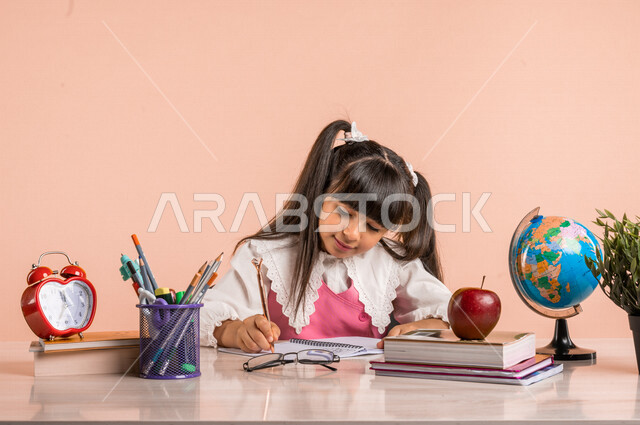 Portrait of a Saudi Arabian Gulf student, in school uniform, sitting at a table, next to a group of pens, textbooks and a globe, doing homework, preparing for the new school year, school education in Saudi Arabia, back to school, colorful background
