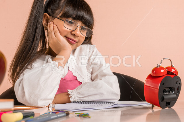 Portrait of a Saudi Arabian Gulf student, in school uniform, sitting at a table, next to a group of textbooks, looking at the small digital clock with gestures indicating thinking, preparing for the new school year, school education in Saudi Arabia, back 