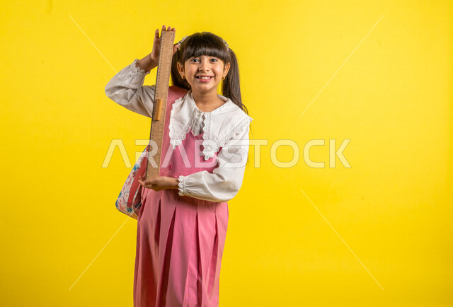 Portrait of a smiling Arab Gulf student, wearing a school uniform, carrying a school bag with a numbered ruler in hand, stationery, preparing for the new school year, school education in Saudi Arabia, yellow background, back to school