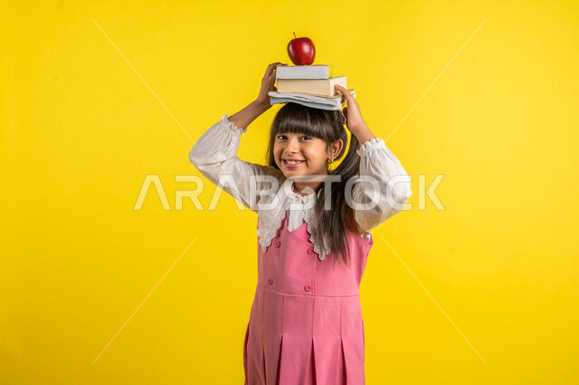 بورتريه لطالبة عربية خليجية مبتمسة، ترتدي الزي المدرسي ، تضع الكتب المدرسية وفاكهة التفاح الأحمر فوق رأسها، الإستعداد للعام الدراسي الجديد، التعليم الدراسي في السعودية، خلفية صفراء، العودة إلى المدارس