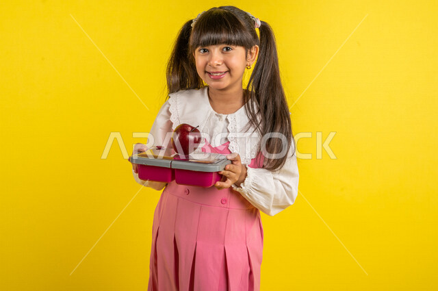 Portrait of a smiling Arab Gulf student, wearing a school uniform, holding a school lunch box and red apple fruit, healthy morning breakfast, preparing for the new school year, school education in Saudi Arabia, back to school, yellow background