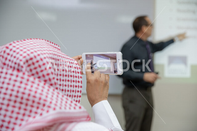 طالب عربي سعودي خليجي في الفصل, يستخدم الهاتف المحمول لتسجيل المحاضرات والدروس, معلم يشرح الدرس في الفصل, مدارس وجامعات, ورش عمل, التعليم في السعوديه