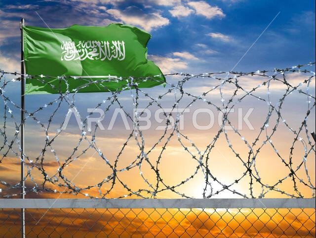 يحمي سياج من الأسلاك الشائكة، على خلفية سماء ملونة، وسارية علم تحمل علم المملكة العربية السعودية، حدود الدولة من المتسللين.