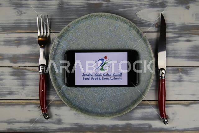 فيرسن، ألمانيا - 9 يونيو 2021: صورة مقرّبة لهاتف ذكي عليه شعار الهيئة العامة للغذاء والدواء السعودية (SFDA) موضوع على طبق مع أدوات مائدة (التركيز على المنتصف).