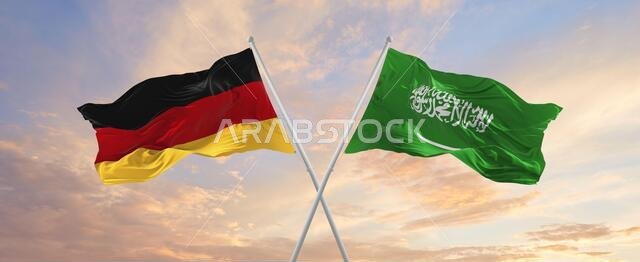 flags of Germany and Saudi Arabia waving in the wind on flagpoles against sky with clouds on sunny day. Symbolizing relationship, dialog between two c