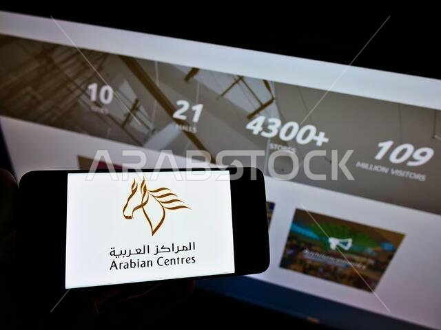 شخص يحمل هاتفًا ذكيًا عليه شعار شركة المراكز العربية (ACC) المشغلة لمركز التسوق، معروضًا على الشاشة أمام صفحة الويب. التركيز على شاشة الهاتف.