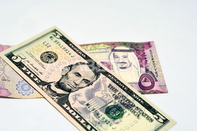 التركيز الانتقائي على ورقة نقدية أمريكية من فئة خمسة دولارات وريال سعودي من فئة خمسة ريالات سعودية، والعملة السعودية والدولار الأمريكي