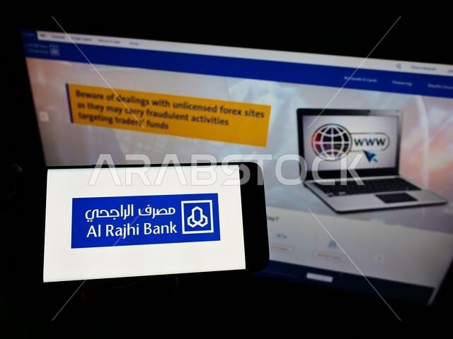 شخص يحمل هاتفًا محمولًا عليه شعار شركة الراجحي المصرفية السعودية على الشاشة أمام موقع الشركة الإلكتروني. التركيز على شاشة الهاتف.