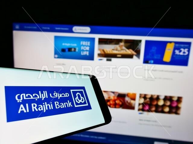 هاتف محمول يحمل شعار شركة الراجحي المصرفية السعودية على شاشته أمام موقعها الإلكتروني. التركيز على منتصف يمين شاشة الهاتف.