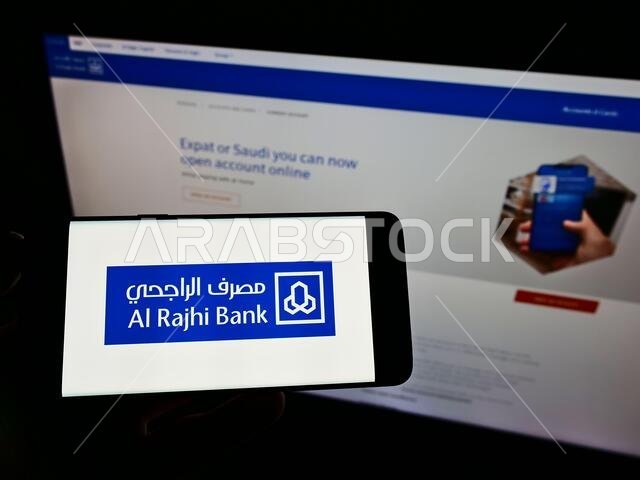 شخص يحمل هاتفًا ذكيًا عليه شعار شركة الراجحي المصرفية السعودية على الشاشة أمام موقع إلكتروني. التركيز على شاشة الهاتف. شخص يحمل هاتفًا ذكيًا عليه شعار شركة الراجحي المصرفية السعودية على الشاشة أمام موقع إلكتروني. التركيز على شاشة الهاتف.