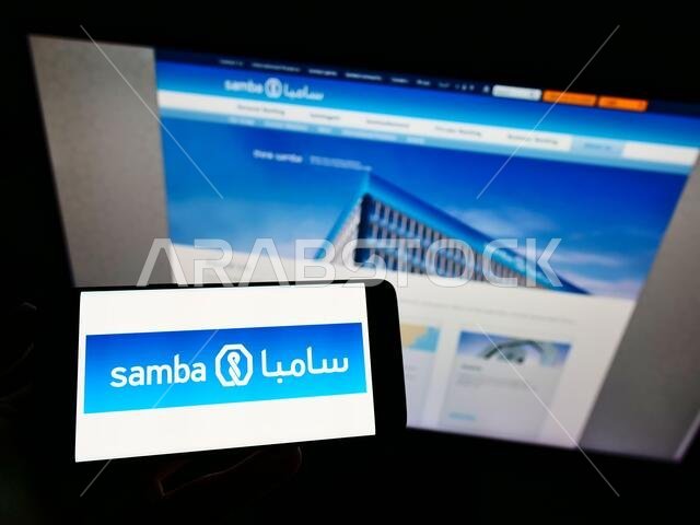 شخص يحمل هاتفًا ذكيًا عليه شعار شركة سامبا المالية السعودية (ش.م.ع.) معروضًا على الشاشة أمام موقع إلكتروني. التركيز على شاشة الهاتف.