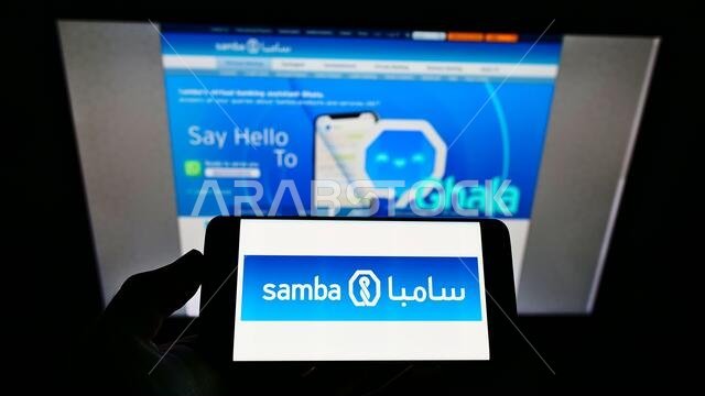 Person holding smartphone with logo of Saudi Arabian company Samba Financial Group SJSC on screen in front of website. Focus on phone display.