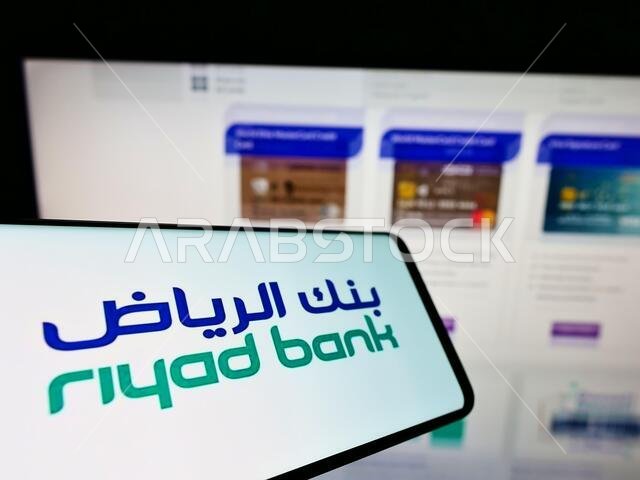 هاتف محمول يحمل شعار شركة الرياض المصرفية السعودية (ش.م.ع) على شاشته أمام موقع الشركة الإلكتروني. التركيز على منتصف شاشة الهاتف.