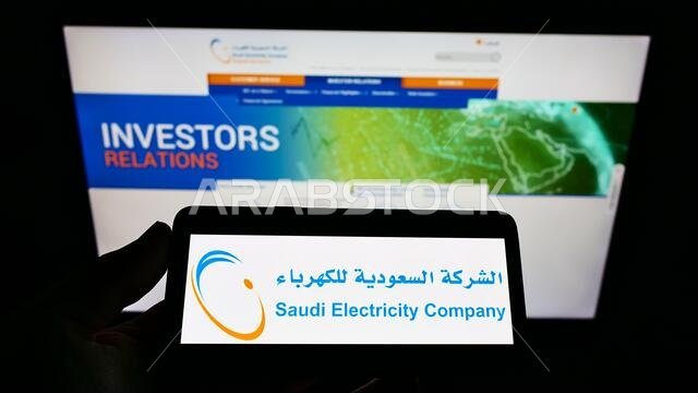 شخص يحمل هاتفًا محمولًا عليه شعار شركة الكهرباء السعودية (SEC) مع صفحة ويب. التركيز على شاشة الهاتف.