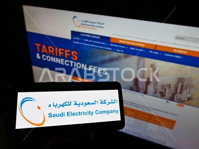 شخص يحمل هاتفًا محمولًا عليه شعار شركة الكهرباء السعودية على الشاشة أمام صفحة ويب. التركيز على شاشة الهاتف.