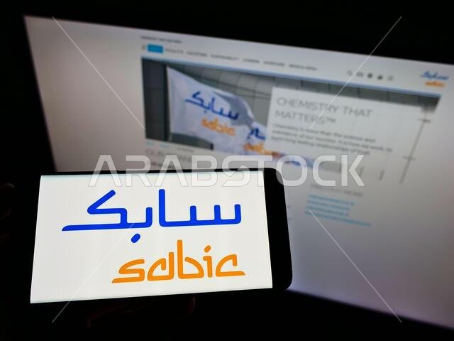 شخص يحمل هاتفًا محمولًا عليه شعار شركة سابك (الشركة السعودية للصناعات الأساسية) على الشاشة أمام صفحة ويب. التركيز على شاشة الهاتف. شخص يحمل هاتفًا محمولًا عليه شعار شركة سابك (الشركة السعودية للصناعات الأساسية) على الشاشة أمام صفحة ويب. التركيز على شاشة الهاتف.