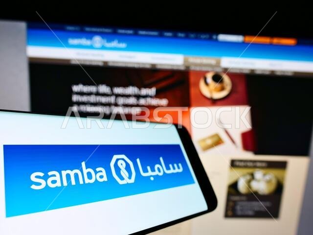 هاتف محمول يحمل شعار شركة سامبا المالية السعودية (ش.م.ع) على الشاشة أمام صفحة الويب. التركيز على منتصف يسار شاشة الهاتف.