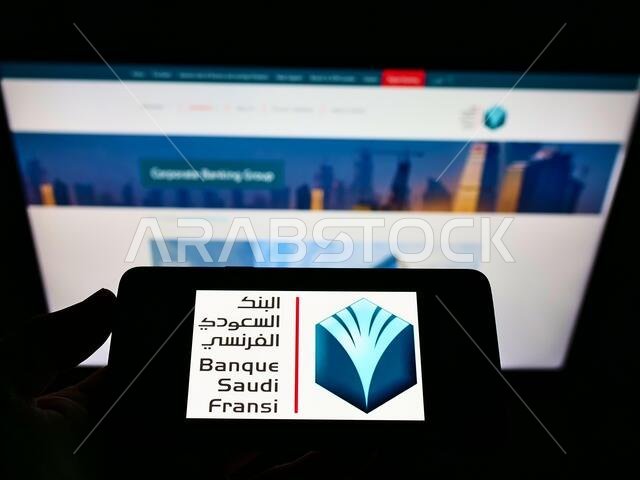 شخص يحمل هاتفًا محمولًا عليه شعار بنك السعودي الفرنسي (BSF) أمام موقع إلكتروني تجاري. التركيز على شاشة الهاتف.