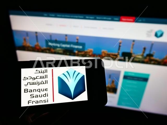 شخص يحمل هاتفًا محمولًا عليه شعار بنك السعودي الفرنسي (BSF) على الشاشة أمام صفحة ويب. التركيز على شاشة الهاتف.