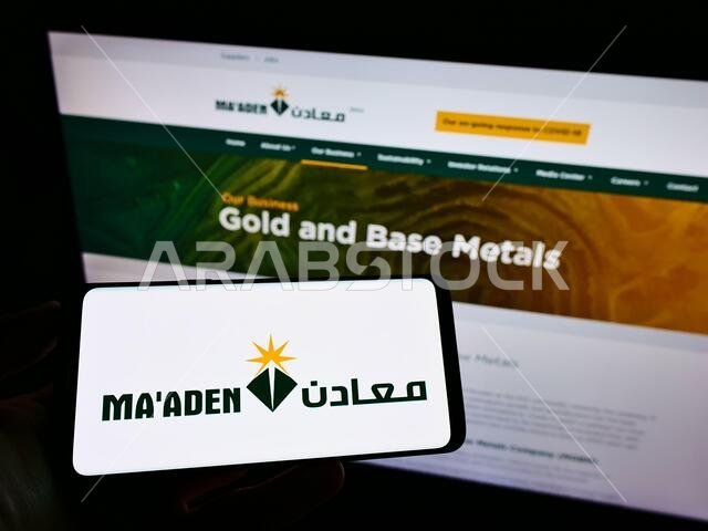 شخص يحمل هاتفًا محمولًا عليه شعار شركة التعدين السعودية (معادن) على الشاشة أمام صفحة الويب الخاصة بالشركة. التركيز على شاشة الهاتف.