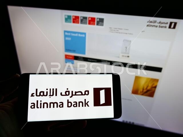 شخص يحمل هاتفًا ذكيًا عليه شعار شركة الإنماء المصرفية السعودية على الشاشة أمام موقع إلكتروني. التركيز على شاشة الهاتف.