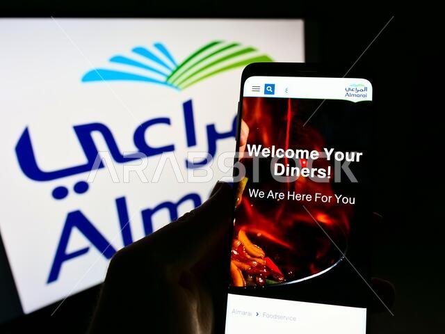 شخص يحمل هاتفًا محمولًا، ويظهر على شاشته موقع شركة المراعي السعودية للألبان، أمام الشعار. التركيز على منتصف شاشة الهاتف.