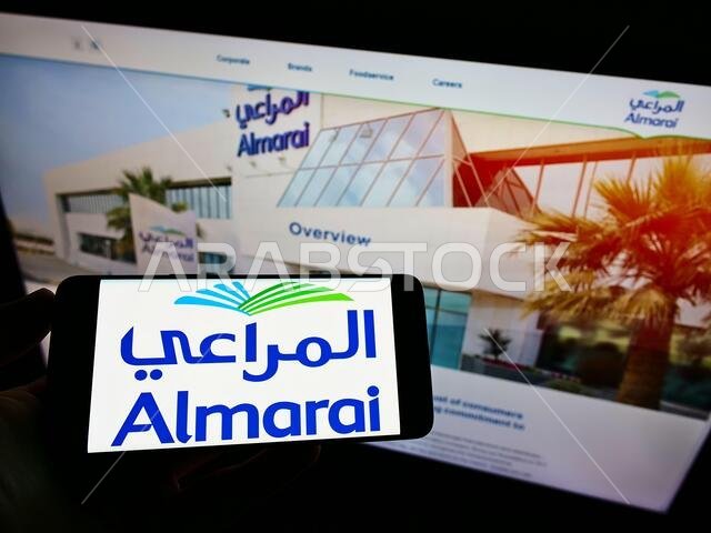 Person holding cellphone with logo of Saudi Arabian dairy company Almarai Company on screen in front of business webpage. Focus on phone display.