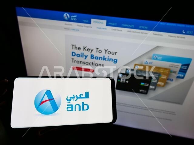 شخص يحمل هاتفًا ذكيًا عليه شعار البنك العربي الوطني السعودي، وهو شركة مالية سعودية، أمام موقع إلكتروني. التركيز على شاشة الهاتف.