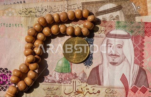 صورة علوية للنقود، الأوراق النقدية والعملات المعدنية للريال السعودي، ومسبحة الصلاة، بتركيز ضحل.